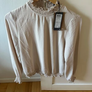 Beige smockad blus från Vero Moda S - Supersöt beige blus från Vero Moda med smockad och rynkad hals samt ärmslut. Långärmad och med lätt rynk vid midjan för en chill och feminin vibe. Materialet är mjukt och har en lätt struktur, perfekt för att lyfta din outfit. 2 knappar bak.