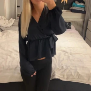 Mörkblå omlott satin blus med knyt - Snygg mörkblå omlottblus med knyt i midjan och volang nedtill. Blusen har långa ärmar och v-ringad hals, vilket ger en cool och trendig look. Perfekt att styla med jeans eller kjol för en chic outfit.