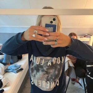 Mörkblå hoodie med månmotiv - Säljer en mörkblå hoodie med coolt tryck av månen och texten 'NOVA LUNA CENERUS' på framsidan. Tröjan har huva med snörning och en stor magficka. Perfekt för dig som gillar rymdinspirerade plagg och vill ha en skön, avslappnad stil.