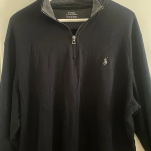 Svart half zip tröja Polo Ralph Lauren - Svart långärmad tröja från Polo Ralph Lauren med half zip och grå insida på kragen. Klassisk broderad logga på bröstet. Tröjan är i mjuk bomull och har en stilren look som funkar till jeans eller chinos. Perfekt för dig som gillar enkel och snygg stil.