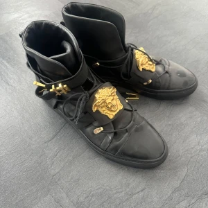 Svarta Versace sneakers med gulddetaljer - Exklusiva svarta high-top sneakers från Versace i skinn med ikonisk Medusa-detalj i guld på ovansidan. Skorna har snörning, platt sula och guldfärgade metalldetaljer vid remmarna. Perfekt för dig som vill sticka ut med lyxig streetstyle.    Mycket dyra vid inköp men används aldrig längre.