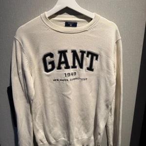 Vit stickad tröja från GANT - Säljer en vit stickad tröja från GANT med broderad logga och texten '1949 New Haven, Connecticut' på bröstet. Tröjan har rund hals, ribbade muddar och lång ärm. Perfekt för dig som gillar klassisk och clean stil.