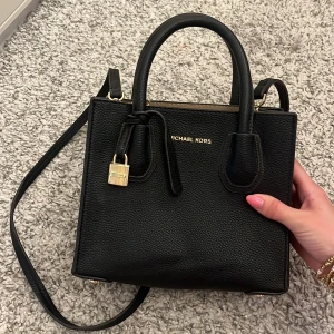 Svart handväska från Michael Kors - Snygg svart handväska från Michael Kors i skinn med guldfärgade detaljer och lås. Väskan har både handtag och axelrem, samt dragkedja upptill. Insidan är beige med guldfärgad logoplatta. Perfekt storlek för det viktigaste och riktigt stilren design. Fick den av min mamma får några år sedan i present. Så har ej, box eller kvitto.