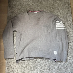 Grå långärmad tröja Thom Browne - Grå långärmad tröja från Thom Browne med klassiska vita ränder på ena ärmen och liten bröstficka. Tröjan har rund hals, knappar i sidan och diskret etikett nertill. Tillverkad i mjuk bomull och har röd, vit och blå detalj i nederkant.
