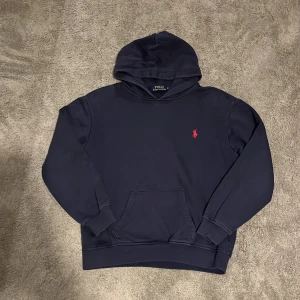 Mörkblå hoodie från Polo Ralph Lauren - Snygg mörkblå hoodie från Polo Ralph Lauren med klassisk röd broderad logga på bröstet. Tröjan har huva, känguruficka och ribbade muddar vid ärmar och nederkant. Strorlek Xs-S Bra kvalité inte jätte använd inga defekter.