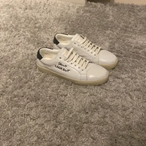 Saint Laurent vita sneakers i skinn - Säljer ett par stilrena vita sneakers från Saint Laurent med svart detalj på hälen och handskriven logga på sidan. Skorna har snörning och platt sula, tillverkade i mjukt skinn för en clean look. Perfekta för dig som gillar minimalistisk design med lyxig touch.   Obs! Ena skon har en defekt där fram.