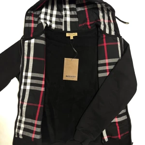 Burburry zip hoodie  - Svart zip hoodie från Burberry med ikoniskt rutigt foder i svart, vitt och rött. Jackan har huva, dragkedja och klassisk streetstil. Perfekt för dig som vill ha en snygg och tidlös look med premiumkänsla. Märkeslapp kvar på jackan. Perfekt for kyliga dagar och kvällar.