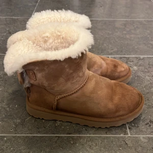 UGG boots med rosett - Säljer ett par klassiska UGG boots i ljusbrun mocka med fluffigt vitt fårskinnsfoder. Baksidan har en söt rosettdetalj och UGG-logga. Storlek: 36