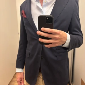 Mörkblå kavaj från Rose & Born - Snygg mörkblå kavaj från Rose & Born, tillverkad i Italien. Klassisk tvåknäppt modell med wide notch lapels, fickor med lock samt en ticket pocket. Kavajen har dubbel slits baktill och är unlined, vilket gör den både lätt och bekväm.  Tyget är ett exklusivt Super 130’s från Reda. Vid ärmsluten finns fyra kissing buttons med öppna knapphål. Mått (liggande, enkel mätning): Armhåla till armhåla: ca 50 cm, Axel till ärmslut: ca 59 cm,	Krage till nederkant: ca 74 cm. Använd men har kvar att ge 😊