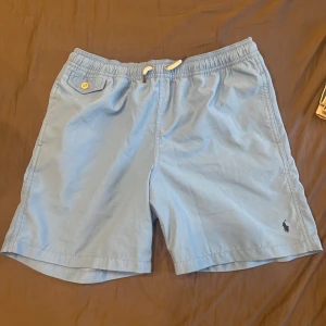Blå badshorts från Polo Ralph Lauren - Snygga ljusblå badshorts från Polo Ralph Lauren med elastisk midja och vit snörning. 