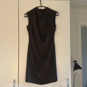 Svart ärmlös Boohoo klänning - Säljer en svart, ärmlös klänning från Boohoo i storlek 36. Klänningen har en snygg draperad halsringning och är tillverkad i ett glansigt satinliknande material. Den är figurnära och har dragkedja i ryggen för enkel påklädning. Perfekt för dig som gillar stilrena och eleganta plagg. Aldrig använd