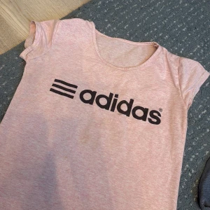 Ljusrosa Adidas t-shirt med tryck - Snygg ljusrosa t-shirt från Adidas med stort svart logotryck på bröstet. Modellen är klassisk med rund hals och korta ärmar. Perfekt för dig som gillar sportiga och stilrena plagg. Materialet känns mjukt och skönt mot huden.