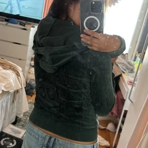 Mörkgrön Juicy Couture hoodie i velour - Säljer en mörkgrön hoodie från Juicy Couture i mjuk velour med glittrigt JC-tryck på ryggen. Tröjan har huva, dragkedja framtill och ribbade muddar. Perfekt för dig som gillar den klassiska Y2K-stilen!