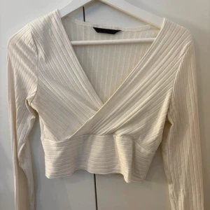 Vit omlottopp med ribbade detaljer - Snygg vit omlottopp med ribbade detaljer och croppad passform. Toppen har lång ärm och djup v-ringning som ger en trendig look. Perfekt att matcha med höga jeans eller kjol för en stilren outfit.