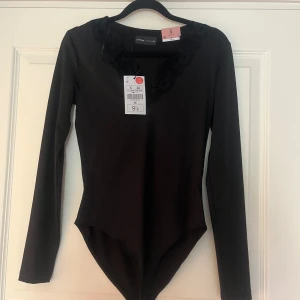 Svart långärmad body från Lefties - Svart body från Lefties i storlek S med lång ärm och snygg spetsdetalj runt halsringningen. Kroppsnära passform som framhäver siluetten och ger en clean look. Perfekt att styla med jeans eller kjol för en trendig vibe.