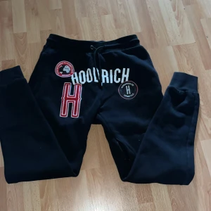 Svarta mjukisbyxor från Hoodrich - Två par svarta mjukisbyxor från Hoodrich med coola tryck och broderade detaljer. Ena paret har stora röda och vita patchar samt snörning i midjan, medan det andra har diskreta loggor och elastiska muddar. Perfekta för chill eller streetwear-stil.