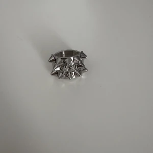 Silvrig ring med nitar - Cool bred ring i silverfärgad metall med flera spetsiga nitar runt hela ringen. Perfekt för dig som gillar edgy och unik stil. Ringen har en chunky design som verkligen sticker ut och ger en tuff vibe till din outfit.