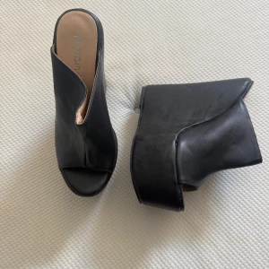 Svarta plattformssandaler  - Använt enstaka gång. Svarta sandaler med hög plattform och öppen tå från Catch. Snygg asymmetrisk design och bred klack ger extra höjd och cool vibe. Tillverkade i skinnimitation med mjuk innersula i beige. Perfekta för dig som gillar statement-skor.