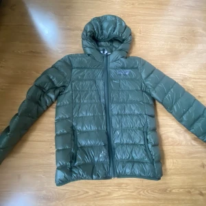 Grön dunjacka EA7 Emporio Armani - Snygg grön dunjacka från EA7 Emporio Armani med huva och dragkedja framtill. Jackan har ett quiltat mönster och logga på bröstet. Perfekt för kalla dagar och har en modern, sportig look som passar till streetwear. Materialet är lätt och syntetiskt. Jackan har några gråa färg fläckar på sig av en penna som säkert går att skrubba bort. Jackan är storlek 150cm och passar att använda på hösten. Jackan kostar 1700kr på zalando där jag köpte den. Hör av er vid frågor eller intresse☺️