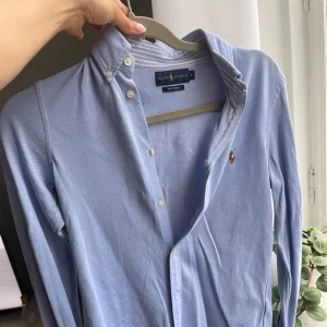 Blå slim fit oxfordskjorta från Ralph Lauren - Klassisk ljusblå oxfordskjorta från Ralph Lauren med broderad logga på bröstet. Skjortan har lång ärm, knappar framtill och button down-krage. Tillverkad i mjuk stretchig bomull som känns skön mot huden. På insidan av ärmarna och kragen syns det vitblå-randiga fina mönstret. 