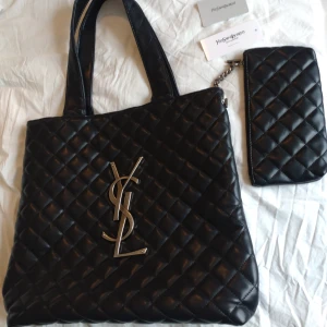 Svart quiltad YSL handväska i skinn - Säljer en helt ny, aldrig använd svart quiltad handväska från Saint Laurent med ikoniskt YSL-emblem i silver framtill. Väskan är i skinn och har två handtag samt en matchande mindre väska med kedjedetalj. Medföljer certifikat kort. Läderflärp med serienummer samt QR kod inuti väskan. Perfekt för dig som vill ha en lyxig och stilren accessoar.kan dock inte garantera äktheten. Kommer från ett pedantiskt hem djur och rökfritt! 