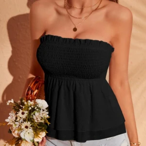 Svart bandeau tubtopp med volang - Supersöt svart bandeau tubtopp med smockad överdel och volang nertill. Toppen är axelbandslös och har en lätt rynkad kant upptill för extra detalj. Perfekt att styla med jeans eller kjol för en chill look.