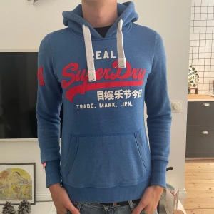 Blå Superdry hoodie med tryck - Snygg blå hoodie från Superdry med stort rött och vitt tryck på bröstet och japanska tecken. Tröjan har huva med vita snören, känguruficka och en röd siffra 4 på ärmen. Perfekt för dig som gillar streetstyle och vill sticka ut. Modellen är 185cm, 75kg