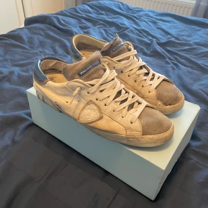 Philippe Model vita sneakers med blå detalj - Snygga sneakers från Philippe Model i vitt skinn och mocka med grå och blå detaljer. Klassisk låg modell med snörning och rund tå. Sulan har tydlig Philippe Model-logga undertill. Perfekta för dig som gillar stilrena och trendiga skor.