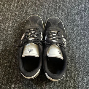 Svarta Adidas sneakers strl 40 - Klassiska svarta Adidas sneakers med vita ränder och vit plös. Skorna har snörning och platt sula, tillverkade i mocka och syntet. Perfekta för dig som gillar en sportig och stilren look. Kostar 150 kr och du måste betala 50 kr för frakt.