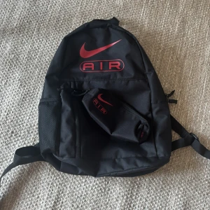 Svart Nike Air ryggsäck med röd logga - Snygg svart ryggsäck från Nike med röd Air-logga framtill. Väskan har flera fack, justerbara axelremmar och en praktisk ytterficka med dragkedja. Tillverkad i slitstark polyester och perfekt för skola, träning eller vardag. Medföljer matchande pennfodral. Denna tvättas och stryks inte innan den skickas. Pris kan diskuteras.