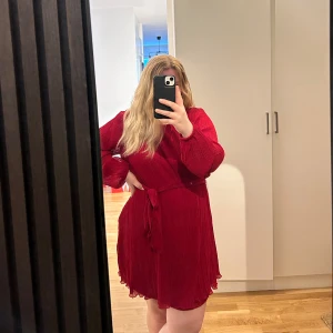 Röd plisserad kortklänning med knyt - Snygg röd kortklänning med plisserat tyg och knytband i midjan. Klänningen har långa ärmar med lätt volang vid ärmslut och en flowy känsla. Perfekt för dig som vill sticka ut med färg och stil. Använd endast två gånger.