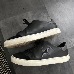 Svarta sneakers från Axel Arigato - Svarta sneakers i skinn från Axel Arigato med broderad fågel på sidan och vit platt sula. Klassisk låg modell med svarta snören och diskret guldfärgad logga. Perfekta för dig som gillar stilrena och trendiga skor. Liten defekt sista bild. BOX TILLKOMMER, NYPRIS 2600