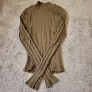 Brun ribbad polotröja - Snygg beige polotröja med ribbat mönster och lång ärm med slits vid handleden. Tröjan har en hög hals och är figurnära i modellen, perfekt att styla med jeans eller kjol. Materialet känns mjukt och stretchigt, vilket gör den bekväm att bära hela dagen.