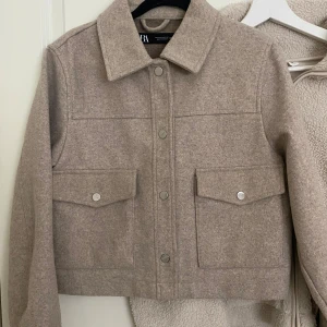 Beige kappa från Zara - Stilren beige kapp från Zara med stora fickor framtill och tryckknappar. Jackan har klassisk krage och är tillverkad i ett mjukt ullblandat material. Perfekt för lager-på-lager och enkel att matcha med det mesta.