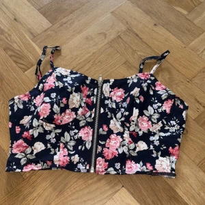 Blommig croppad topp från SHEIN - Supersöt croppad topp från SHEIN med blommigt mönster i rosa, beige och vitt på mörkblå botten. Toppen har smala axelband, markerade kupor och dragkedja framtill som ger en edgy vibe. Perfekt för sommarens alla häng och outfits!