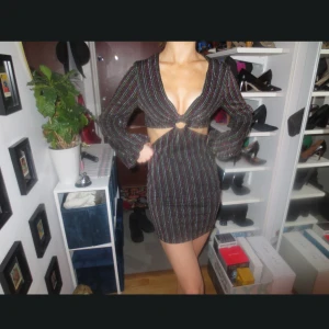 Glittrig svart kortklänning med cut out - Supersnygg svart kortklänning med glittriga ränder i flera färger. Djup v-ringning, långärmad och med cut out-detalj framtill samt ring i midjan. ✨✨🪩 Ny utan prislapp