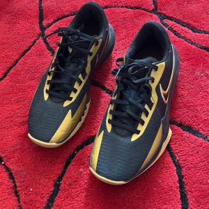 Svarta och guldiga Nike sneakers - Snygga svarta Nike sneakers med guldiga detaljer och klassisk swoosh på sidan. Skorna har snörning och en sportig design med platt sula, perfekta för dig som gillar att sticka ut med stilrena färgkombinationer.