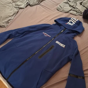 Blå Hoodrich vindjacka med huva M - Blå vindjacka från Hoodrich med tryck på bröstet, ryggen och huvan. Jackan har hel dragkedja, huva och en bröstficka med dragkedja. Snygga svarta detaljer på ärmen och sportig streetkänsla. Perfekt för dig som gillar urban stil. Otroligt fin tröjan super bra skick