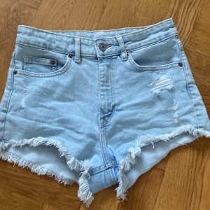 Ljusblåa jeansshorts från Divided stl 38 - Snygga ljusblåa jeansshorts från Divided med hög midja, slitna detaljer och fransig kant. Klassisk femficksmodell och bälteshällor. Perfekta för varma sommardagar och ger en avslappnad vibe.