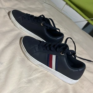 Mörkblå sneakers från Tommy Hilfiger - Snygga mörkblå sneakers från Tommy Hilfiger med klassiska ränder i rött, vitt och blått på sidan. Skorna har vita sulor, rund tå och snörning. Tillverkade i skinnliknande material och har diskret logga på plösen. Perfekta för en stilren och sportig look.