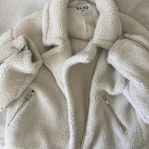 Beige teddyjacka från NA-KD, stl 40 - Supermjuk och fluffig teddyjacka från NA-KD i beige. Jackan har stora kragar, dragkedjor på fickor och ärmar samt en oversized känsla. Perfekt för kalla dagar när du vill vara både varm och snygg. Passar dig som gillar en chill och trendig stil.