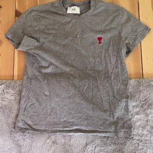 Grå t-shirt från Ami med rött hjärta - Snygg grå t-shirt från Ami Alexandre Mattiussi med klassisk passform och korta ärmar. På bröstet finns en broderad röd hjärtlogga. Tillverkad i mjuk bomull som känns skön mot huden. Perfekt för en clean och stilren look.