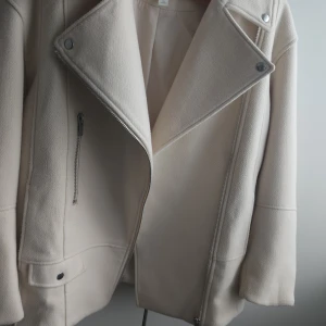Beige jacka H&M - Snygg beige jacka i ullmix med bred krage och silvriga dragkedjor. Jackan har en klassisk bikerstil med snedställd dragkedja, fickor med dragkedja och tryckknappar på kragen. Perfekt för dig som gillar stilren och trendig look.