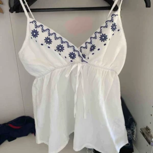 Vit linnetopp med knyt - Supersöt vit linnetopp med tunna axelband och blå blommor längs v-ringningen. Toppen har knyt i fram. linnet är inte så stretchigt men jätte gullig. sålänge man inte har svart bh så är den inte genomskinlig 