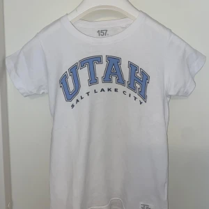 Vit t-shirt UTAH 157 Salt Lake City - Vit t-shirt från lager 157 med blå text 'UTAH Salt Lake City' på bröstet. Klassisk passform med rund hals och korta ärmar. Perfekt för dig som gillar college-vibe och enkel stil.