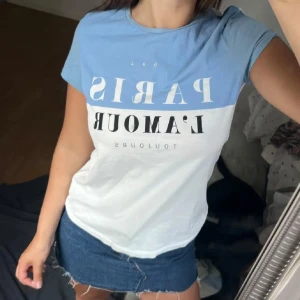 Blåvit t-shirt med Paris-tryck - T-shirt i tvåfärgad design med ljusblå upptill och vit nedtill. Stor text med 'PARIS L'AMOUR' framtill. Klassisk rund halsringning och korta ärmar. Perfekt för en chill och trendig look.