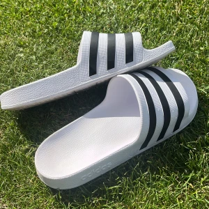 Vita Adidas sandaler med svarta ränder - Fräscha vita Adidas sandaler med klassiska svarta ränder över foten. Tillverkade i syntetmaterial med räfflad sula för extra grepp. Perfekta för chill dagar eller häng vid poolen. Enkel slip-on-modell med Adidas-logga på sidan. lite orangerosa undertill pga använt dom två ggr max. pris kan diskuteras.