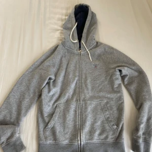 Grå hoodie med dragkedja från Gant - Snygg grå hoodie från Gant i storlek XS med dragkedja och huva med vita snören. Tröjan har två stora fickor framtill och broderad Gant-logga på bröstet. Perfekt för en chill och sportig look.
