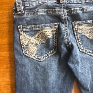 Blå bootcut jeans med broderade fickor - Snygga blå jeans med bootcut-modell och broderade bakfickor i beige och silver. Jeansen har klassisk femficksdesign, markerade sömmar och är tillverkade i mjukt denimtyg. Perfekta för dig som gillar detaljer och en lite vidare passform nertill.