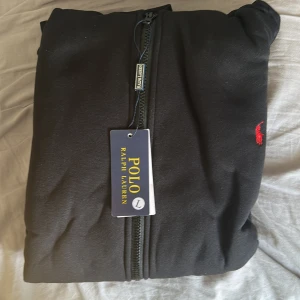 Svart zipjacka från Polo Ralph Lauren - Svart zipjacka från Polo Ralph Lauren med klassisk röd broderad logga på bröstet. Jackan har dragkedja hela vägen, hög krage och praktiska fickor framtill, perfekt för en chill och stilren look.